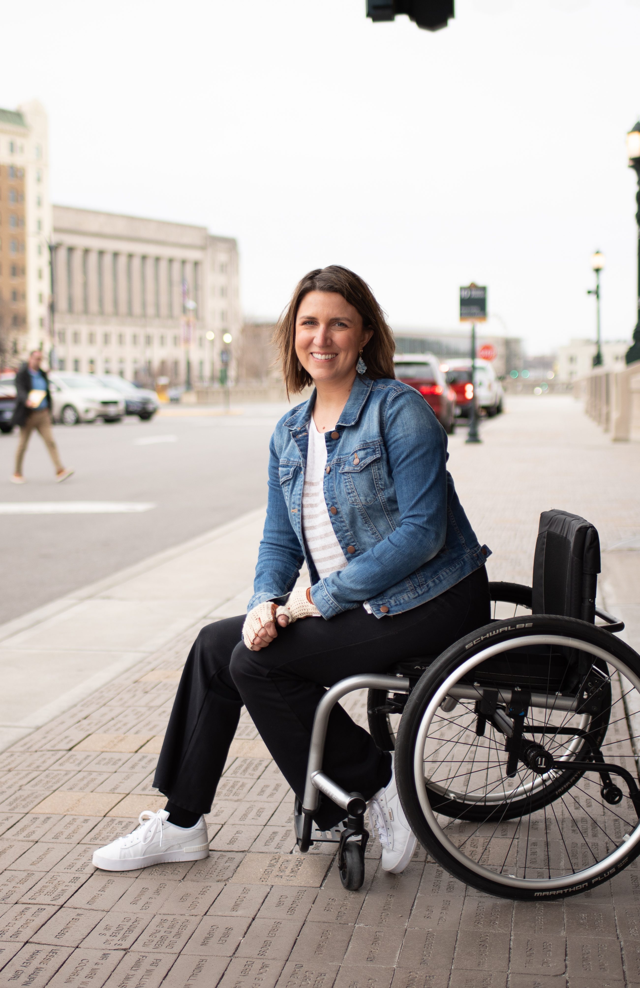 Stephanie sits on her wheelchair, leaning forward. She is outdoors, wearing a jean jacket.